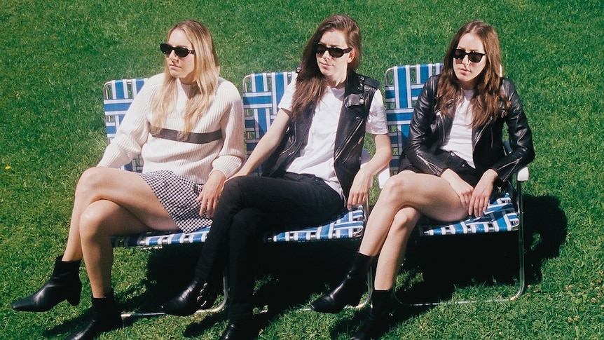 The three Haim sisters sit in blue deckchairs wearing sunglasses and crossing their legs