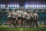Fijian rugby team hudle together, white tee and black shorts uniform, in stadium grounds under bright lights.