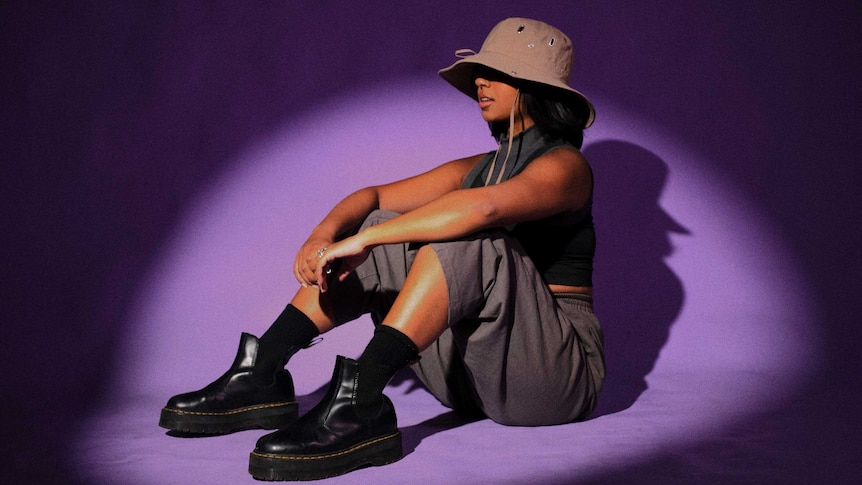 Munasib sits under a spotlight against a purple backdrop. She wears a beige hat, grey singlet & pants & black docs.