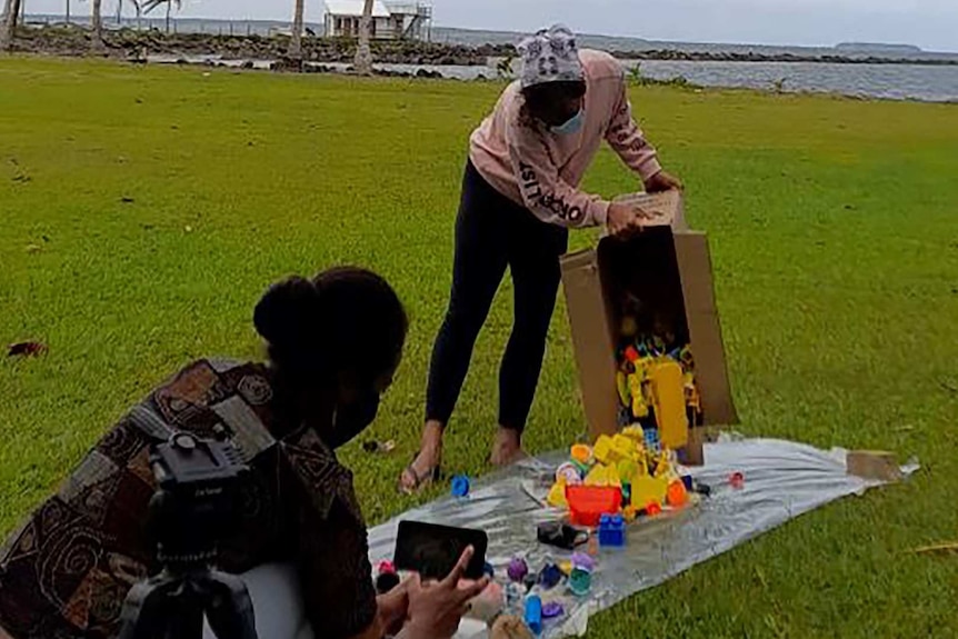 A woman empties plastic rubbish from a tub on a mat on some lawn by the coast while another woman films it on a phone.