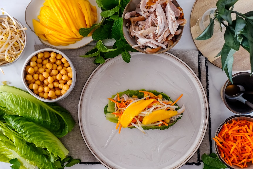 A rice paper wrapper on a plate with fresh veggies, chicken, mango and herbs, ready to roll.