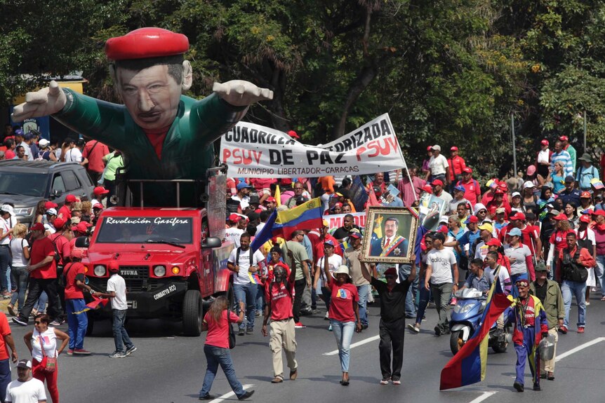 A giant blow-up figure of Venezuelan President Nicolas Maduro driven in a red car alongside people in red walking on the road.