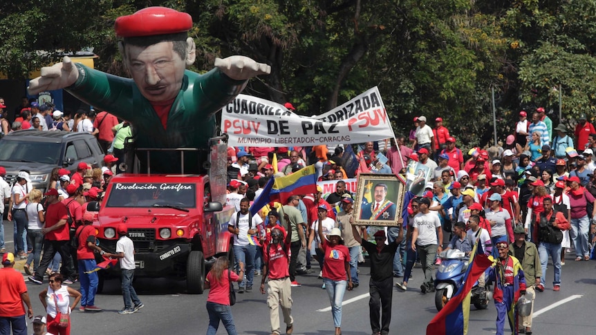 A giant blow-up figure of Venezuelan President Nicolas Maduro driven in a red car alongside people in red walking on the road.