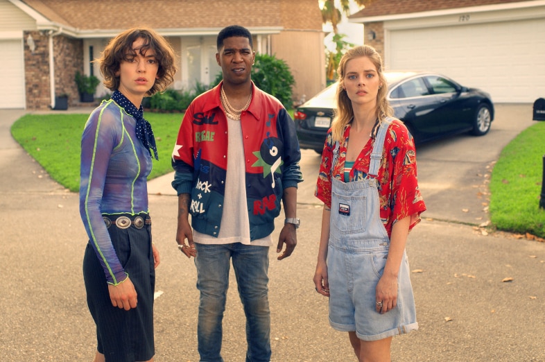 A brunette woman, man with buzz fade and blonde woman in 80s attire stand in sunny suburban cul de sac with worried expressions.