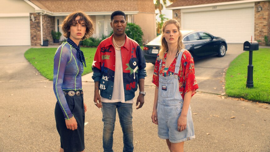 A brunette woman, man with buzz fade and blonde woman in 80s attire stand in sunny suburban cul de sac with worried expressions.
