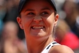Ash Barty makes a fist with one hand and holds a racket and as she looks away from the camera with a grin on her face.