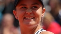 Ash Barty makes a fist with one hand and holds a racket and as she looks away from the camera with a grin on her face.