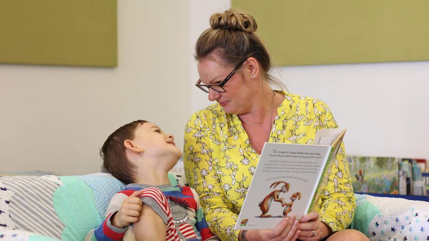 A woman reads a book to a young boy, they are making silly faces at each other.