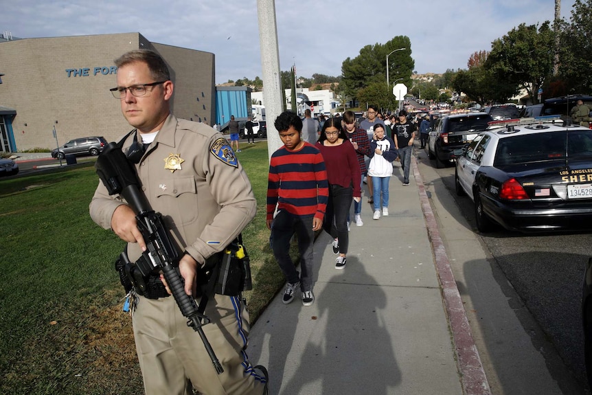 A policeman carries a gun as he walks in front of a line of students.
