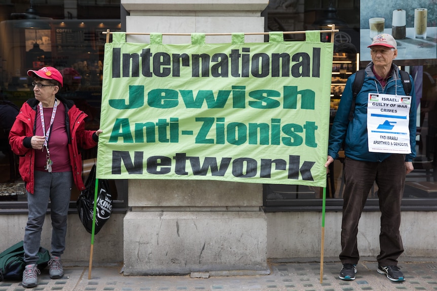 Older man and woman holding sign that reads 'International Jewish Anti-Zionist Network'.