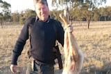 A grazier holds up a dead wild dog, with the pistol he used to shoot it visible in its underarm holster