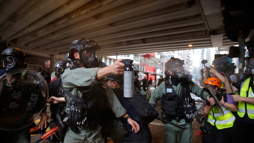 a police officer in a mask holds out a can of pepper spray and sprays it at protesters as media with cameras watch