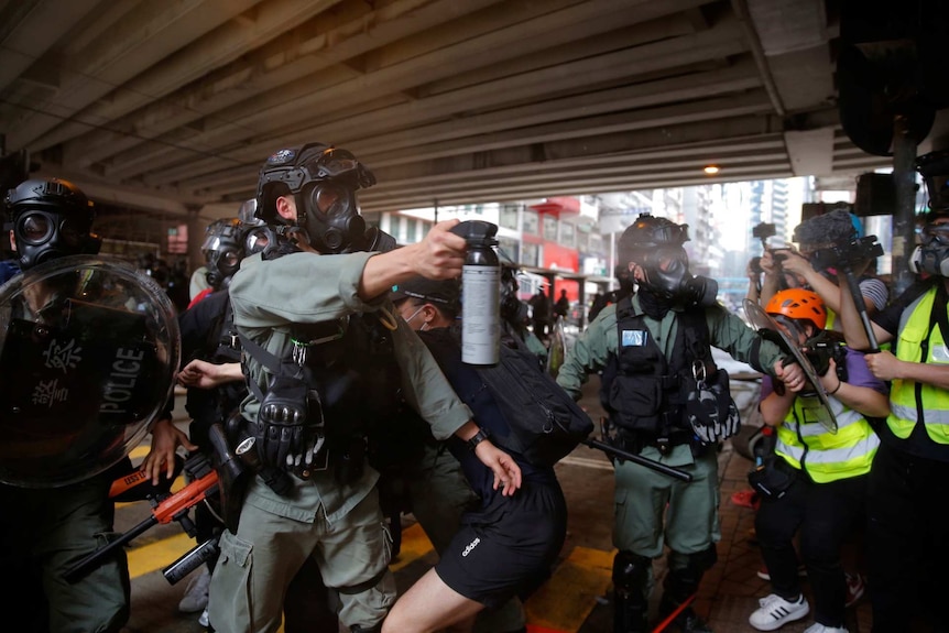 a police officer in a mask holds out a can of pepper spray and sprays it at protesters as media with cameras watch