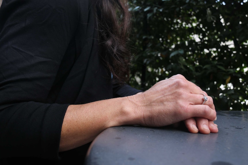 A pair of hands rest folded one on top of the other. One has a ring on the ring finger.