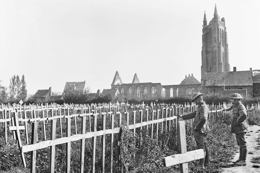 Two soldiers in uniform inspect wooden crosses lining a field outside a church building.