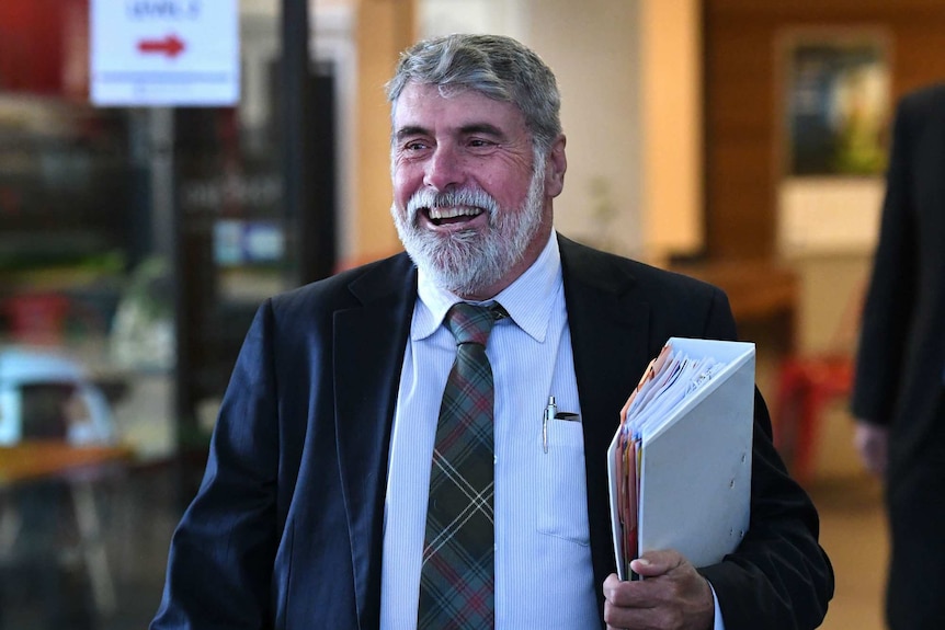 A smiling man in a suit walks down the street carrying a folder.