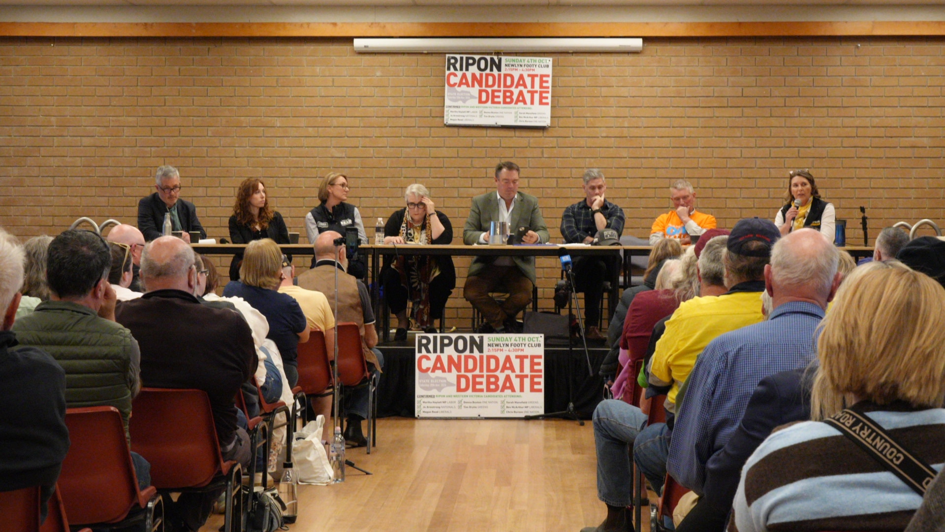 Eight men and woman sit at a long table in front of a crowd in a community hall.