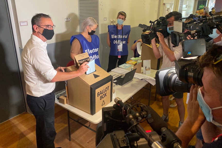 A man puts his ballot into a box while two voting officials look on, along with TV cameras