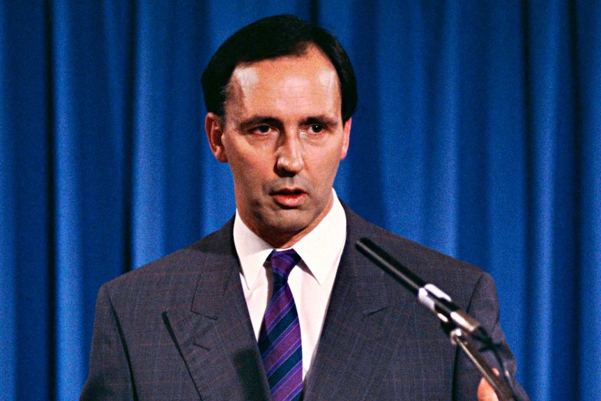 Man in a dark suit stands at a lecture and addresses the crowd 