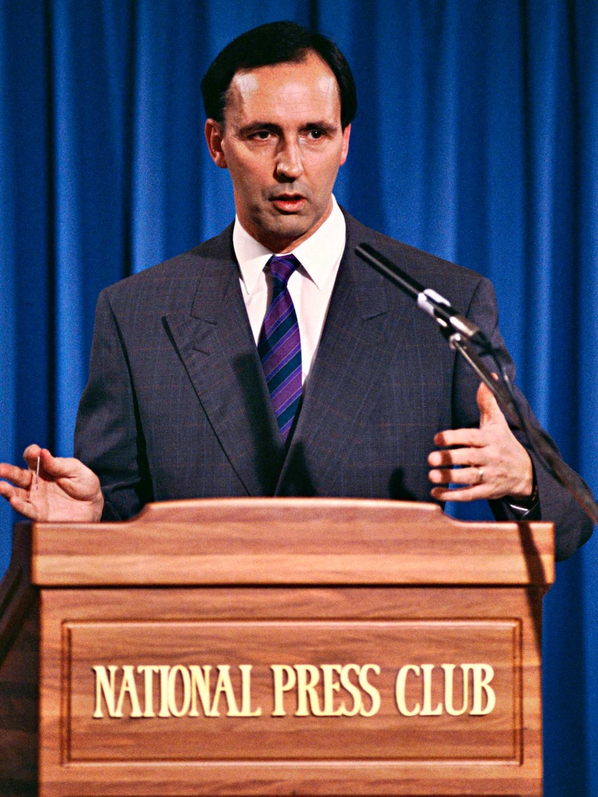 Man in a dark suit stands at a lecture and addresses the crowd 