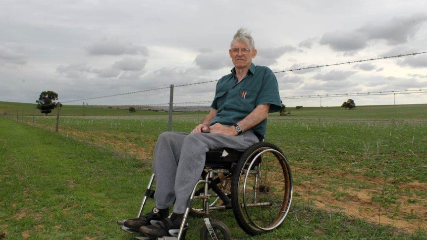 Ian Minty sits in a grassy paddock in a wheel chair. Behind him a wire fence stretches into the distance.