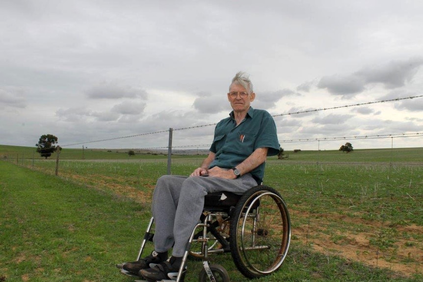 Ian Minty sits in a grassy paddock in a wheel chair. Behind him a wire fence stretches into the distance.