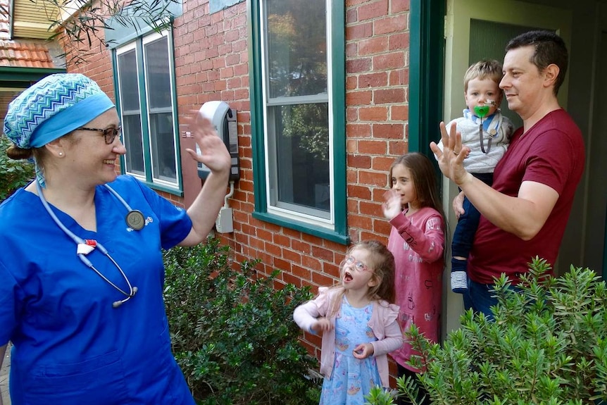 A doctor wearing blue scrubs waves goodbye to her husband and three children at the door of their home.