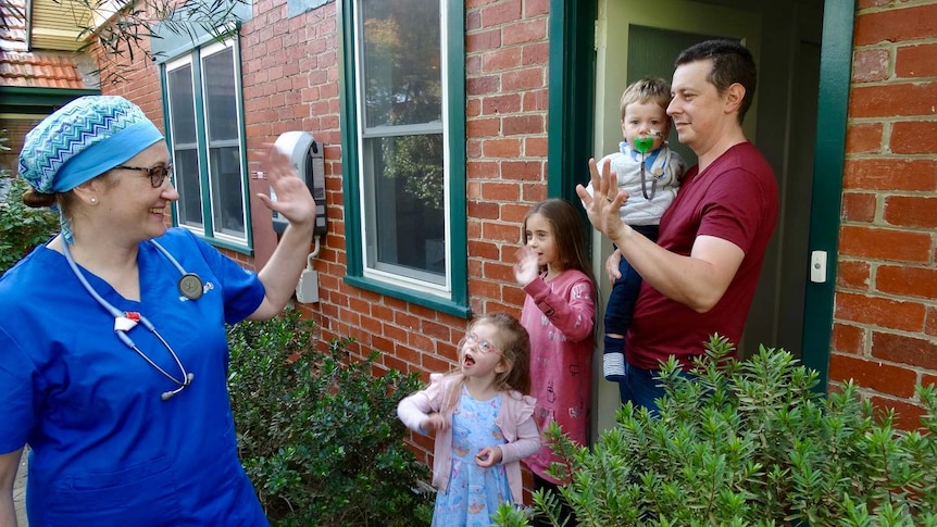 A doctor wearing blue scrubs waves goodbye to her husband and three children at the door of their home.