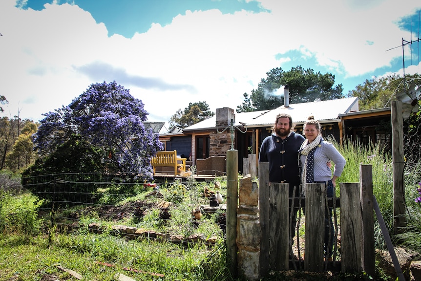 A man and woman standing outside a house with chickens behind a fence.