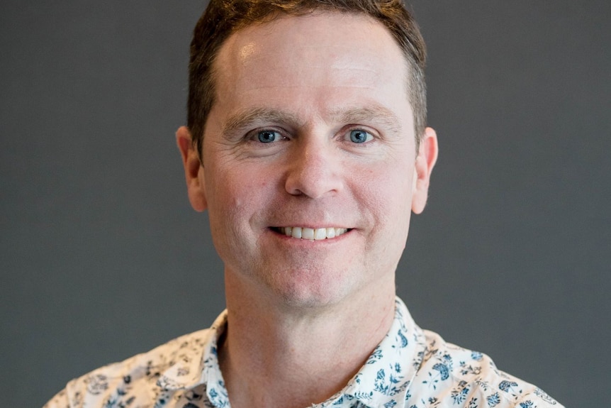 A white man with blue eyes and short brown hair stands smiling in front of a grey wall.