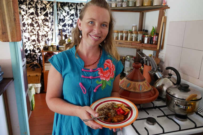 Woman standing in her kitchen holding a bowl of food she'd prepared with Quinoa and Moroccan style tagine.