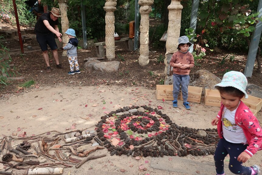 Three children and a teacher run around in a back yard where children have made a large snail out of rocks, sticks and flowers.