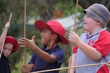 School students in uniforms and red hats hold wooden spears, trees behind.