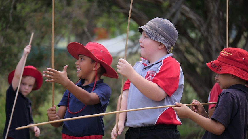 School students in uniforms and red hats hold wooden spears, trees behind.