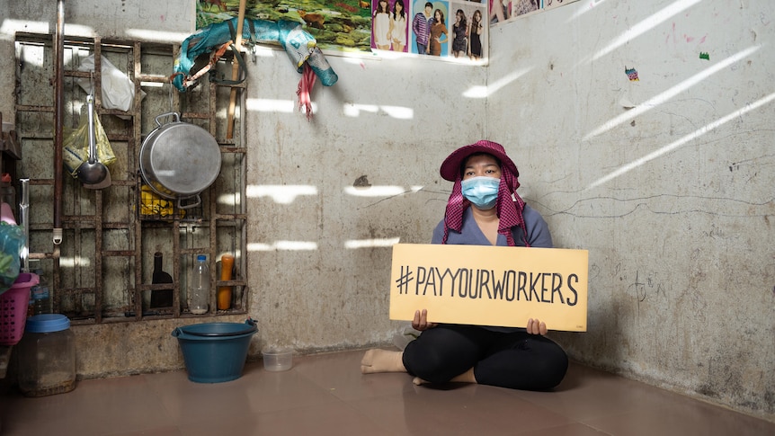 A Cambodian woman sitting in mask and hat holding sign reading "pay your workers".