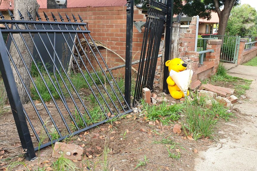 A teddy with flowers is left outside a knocked down gate on a suburban street