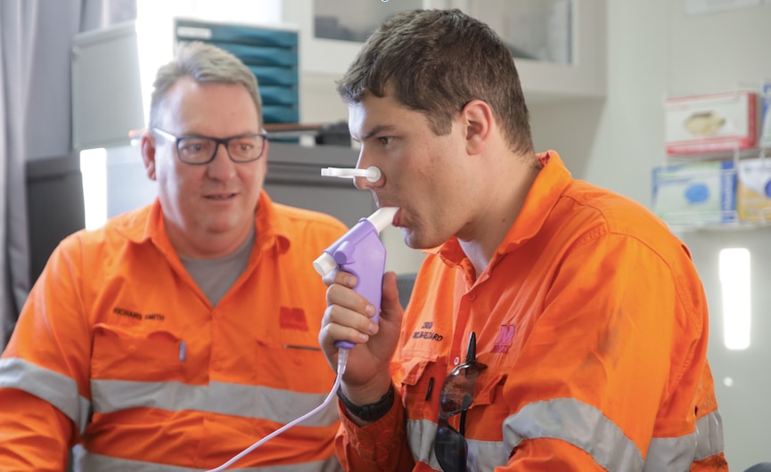 Dois homens com roupas de trabalho laranja de alta visibilidade, um dos quais respira através de um aparelho roxo com clipe nasal. 