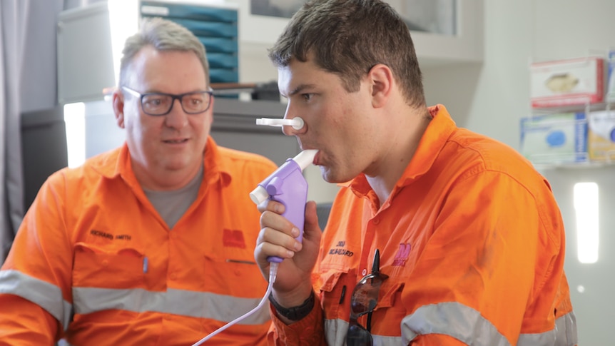 Two men in orange hi-vis workwear, one of whom is breathing into a purple device with a clip on his nose. 