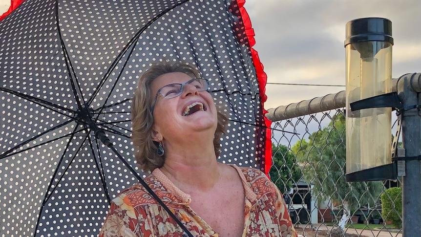 A woman with glasses has a big smile, holding an umbrella next to a rain gauge.