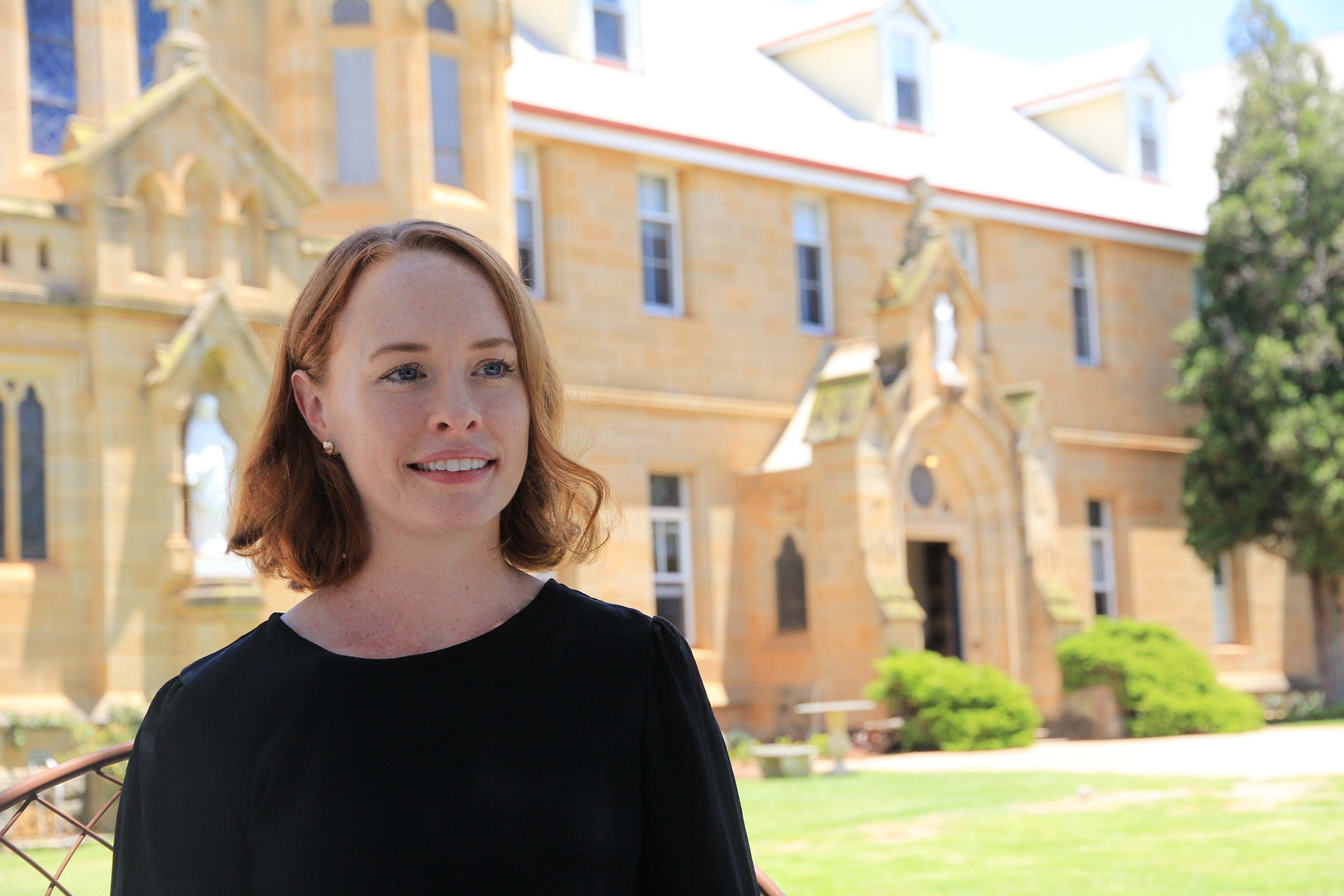 Женщина с короткими каштановыми волосами стоит возле большого особняка из песчаника.