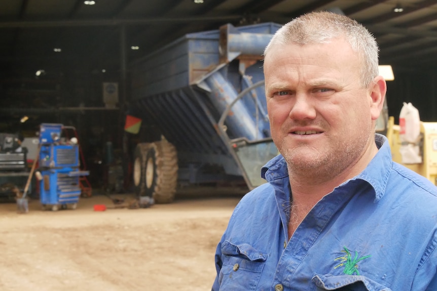 A man with short grey hair stands in front of a machinery shed on a farm.