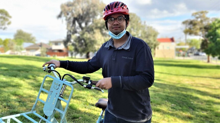 Raiyan stands in a park holding up a blue bike, he wears glasses and a red helmet.