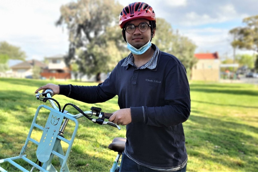 Raiyan stands in a park holding up a blue bike, he wears glasses and a red helmet.
