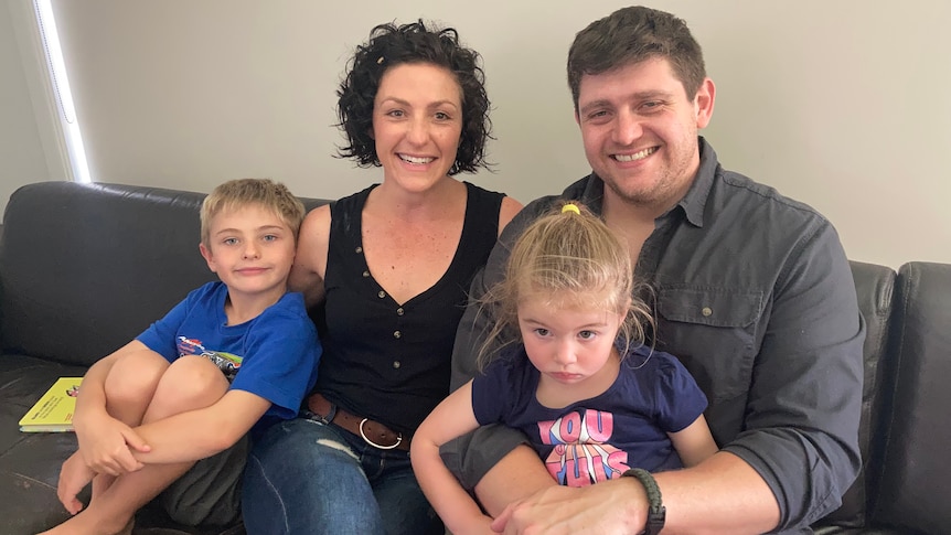 A smiling woman with short, black curly hair, and a black top and jeans sits with a man and a two young children on a sofa.