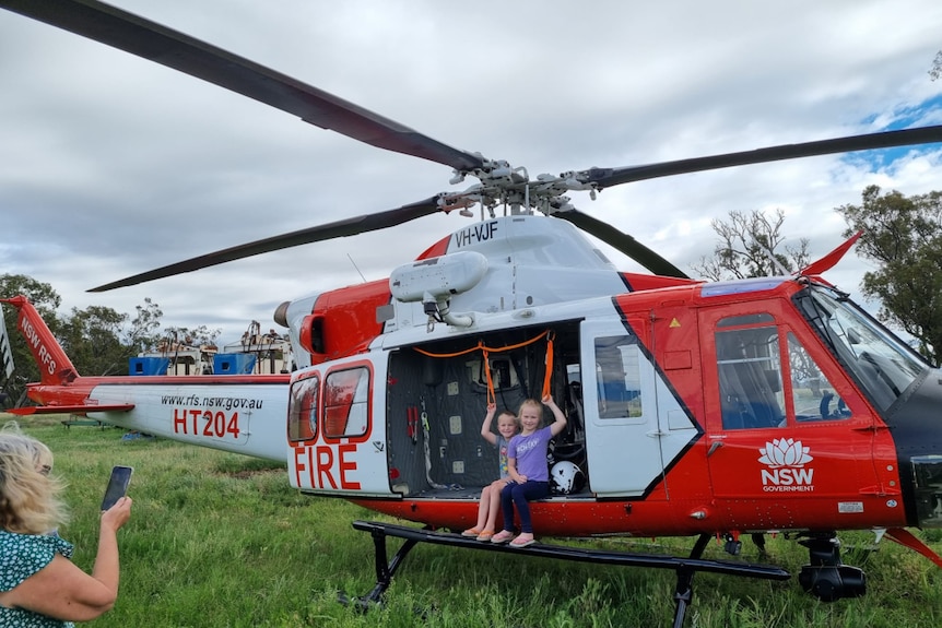 Two children, smiling, sit on the opening to a red and white helicopter