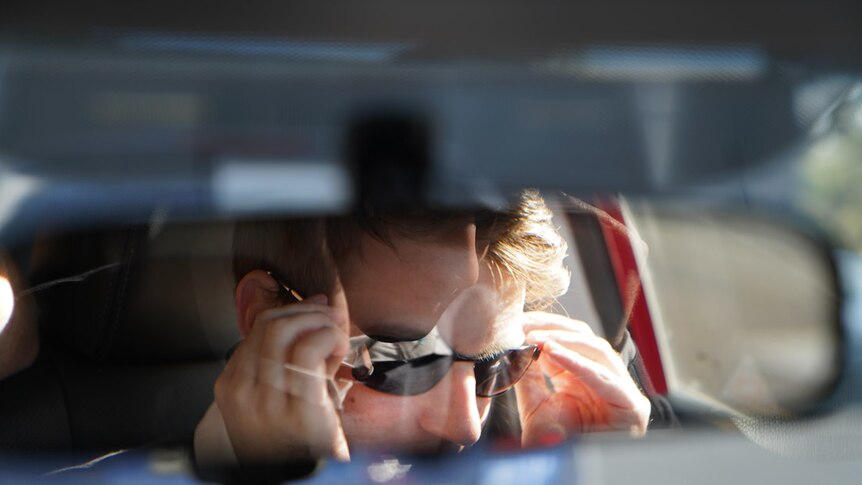 A car's rear view mirror reflects a man putting sunglasses on his face.