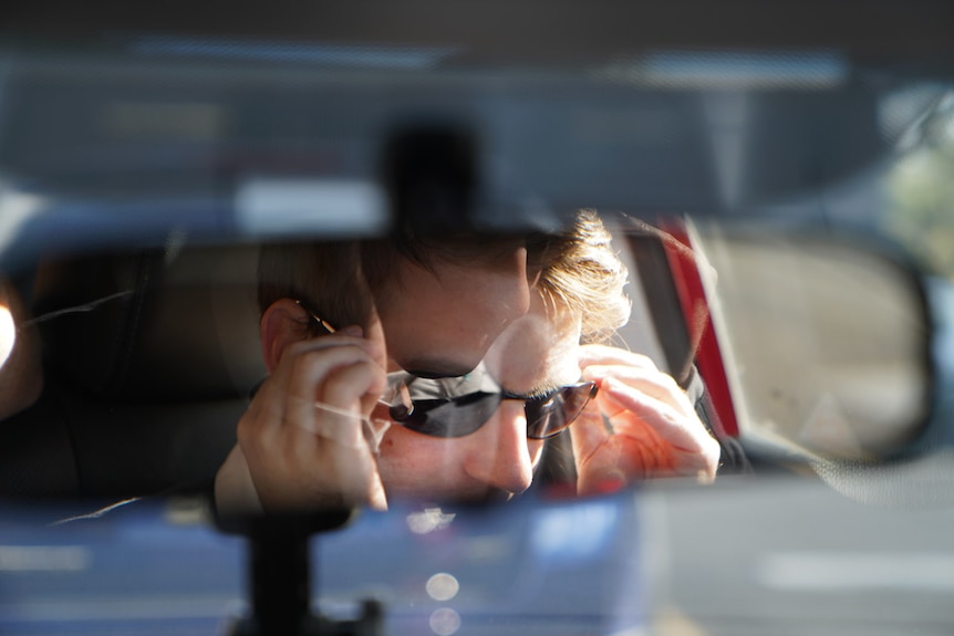 A car's rear view mirror reflects a man putting sunglasses on his face.