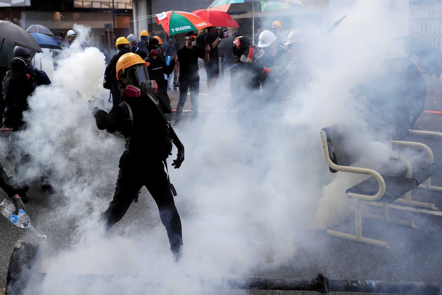 Surrounded by protesters and tear gas, a person wearing a hard-hat and gas mask, throws a gas cannister.