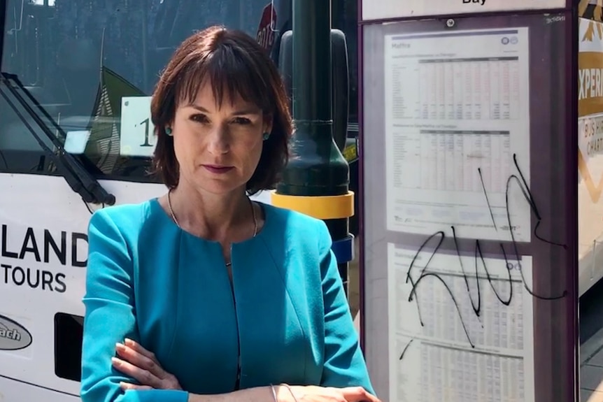 a woman with brown hair wearing a blue jacket stands in front of a bus and timetable. 