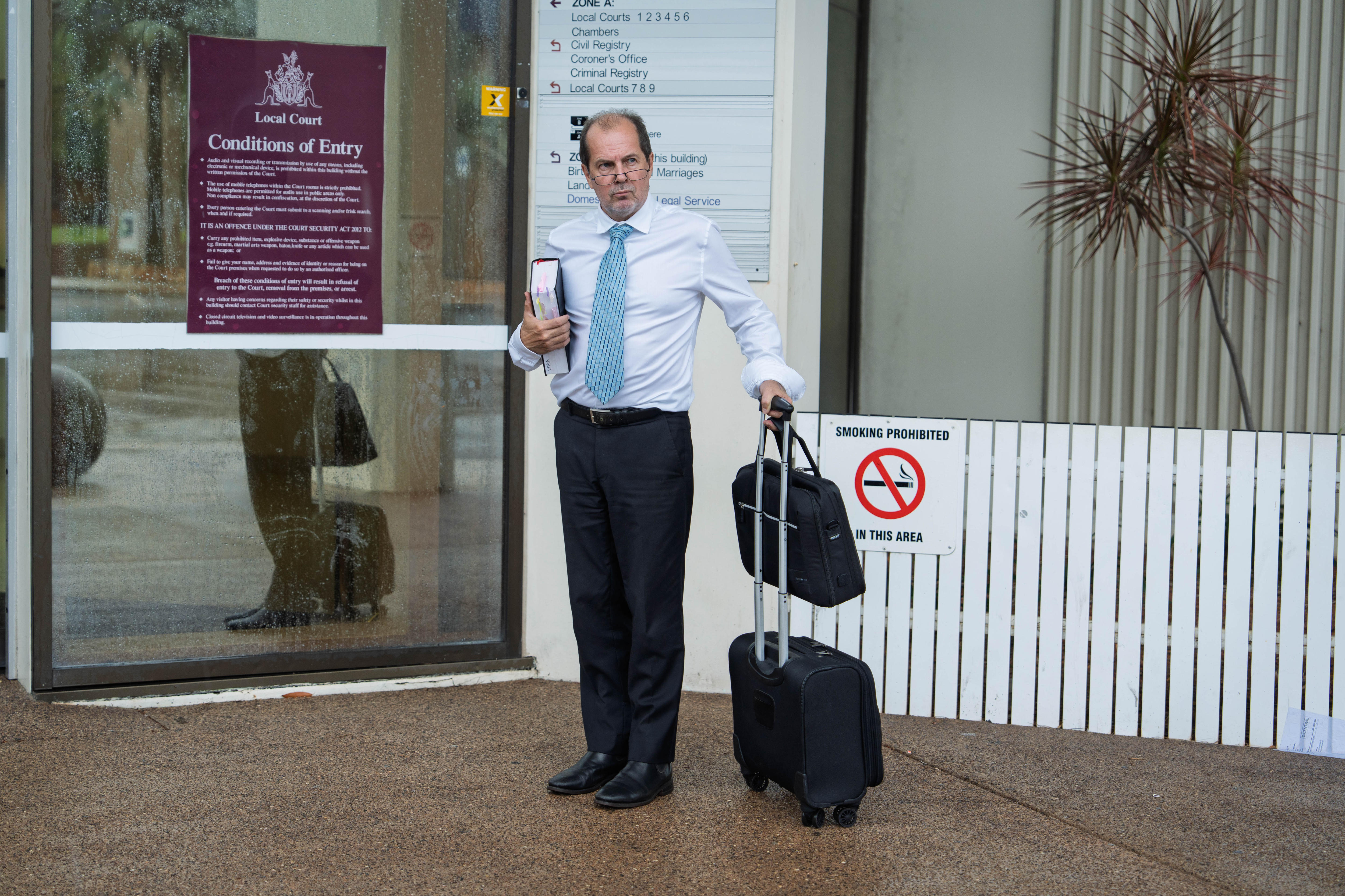 Um homem de camisa branca, calça preta, segurando uma mala, parado em frente à quadra enquanto chove. Gravata azul, expressão séria.